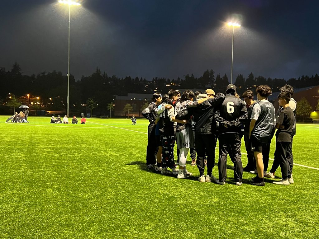 a group of young men huddle up on the turf field under the lights